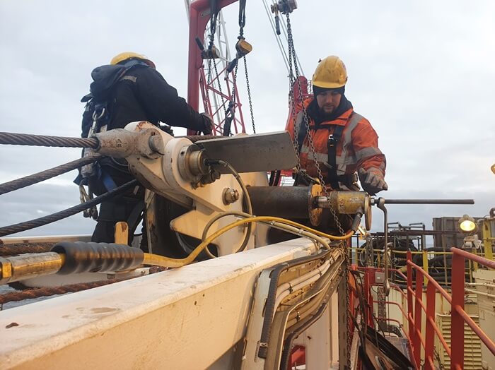 Na zdjęciu widzimy dwóch pracowników wykonujących zadania techniczne na terenie przemysłowym — prawdopodobnie na platformie offshore lub w zakładzie energetycznym. Ubrani są w odzież ochronną: kaski, uprzęże i odblaskowe kombinezony, co podkreśla przestrzeganie wysokich standardów BHP. Otaczają ich elementy maszyn — w tym wyciągarki, liny i komponenty hydrauliczne.