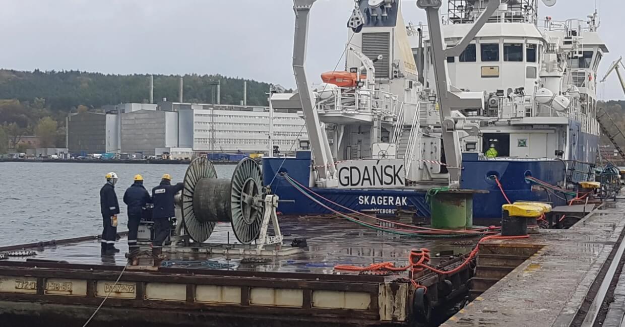 Zdjęcie przedstawia statek, na którym widnieją napisy "GDANSK" i "SKAGERAK". Jest on wyposażony w różne urządzenia pokładowe, takie jak dźwigi, liny i bębny. Widać, że jego rufa jest połączona z pomostem. Na pomostach pracuje grupa trzech pracowników w odblaskowych kurtkach i żółtych kaskach. Stoją oni wokół dużej, metalowej szpuli z grubą liną lub kablem. Ich pozy sugerują, że są zajęci manewrowaniem lub mocowaniem lin.