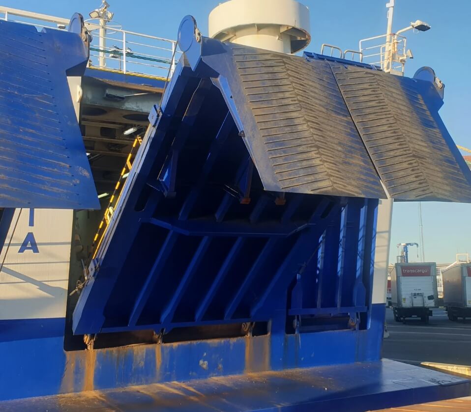 Na zdjęciu widoczna jest podniesiona rampa statku typu Ro-Ro, przeznaczonego do transportu pojazdów i ładunków. Rampa ma niebieską, antypoślizgową powierzchnię i aktualnie znajduje się w pozycji uniesionej, co sugeruje przerwę w załadunku lub rozładunku. W tle widać ciężarówki oraz infrastrukturę portową, co wskazuje na komercyjny terminal morski.