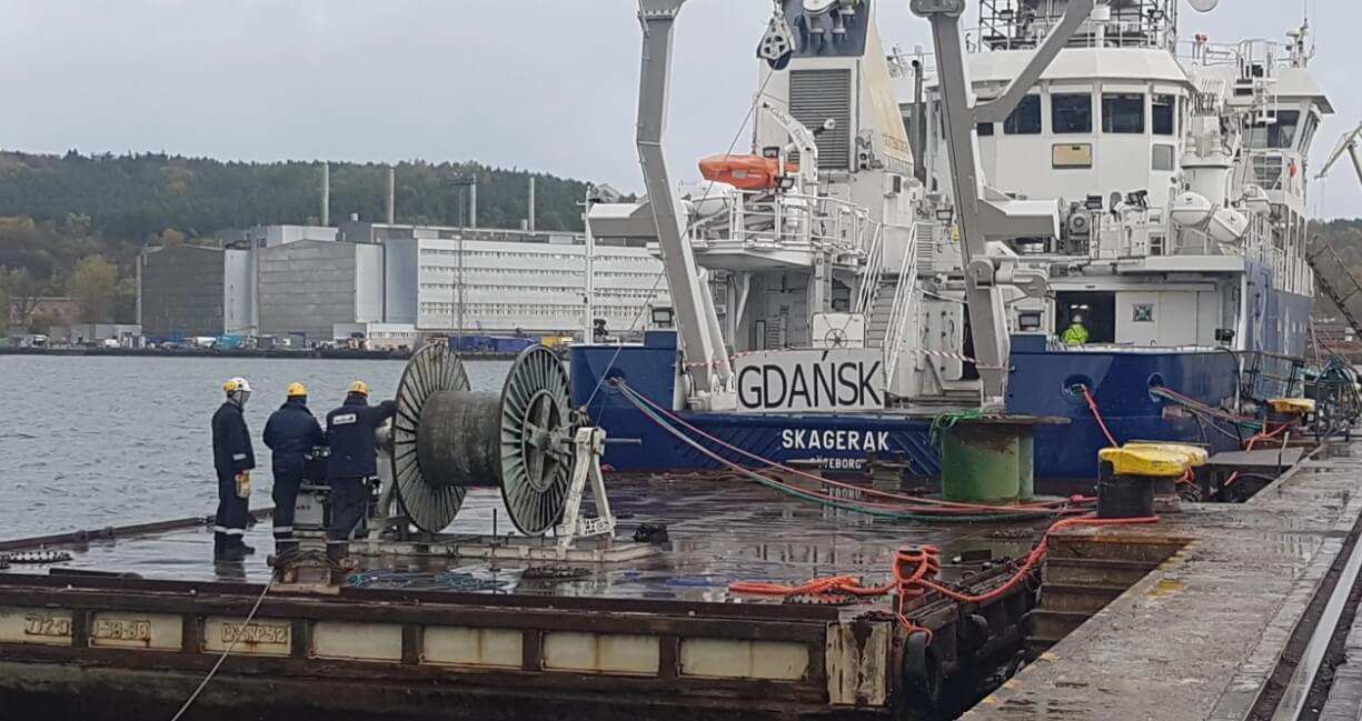 Na zdjęciu widzimy nabrzeże portowe w Gdańsku, gdzie zacumowany jest statek o nazwie „SKAGERAK” — prawdopodobnie jednostka badawcza lub techniczna, wyposażona w dźwigi i sprzęt morski. Na pierwszym planie trzech pracowników w kaskach i odzieży ochronnej stoi obok dużych bębnów z kablami, co sugeruje trwające prace serwisowe lub przygotowania do operacji. Mokra nawierzchnia odbija światło, wskazując na niedawny deszcz. W tle widoczne są budynki przemysłowe oraz zalesiony obszar, co tworzy charakterystyczny krajobraz nadmorskiej strefy przemysłowej.