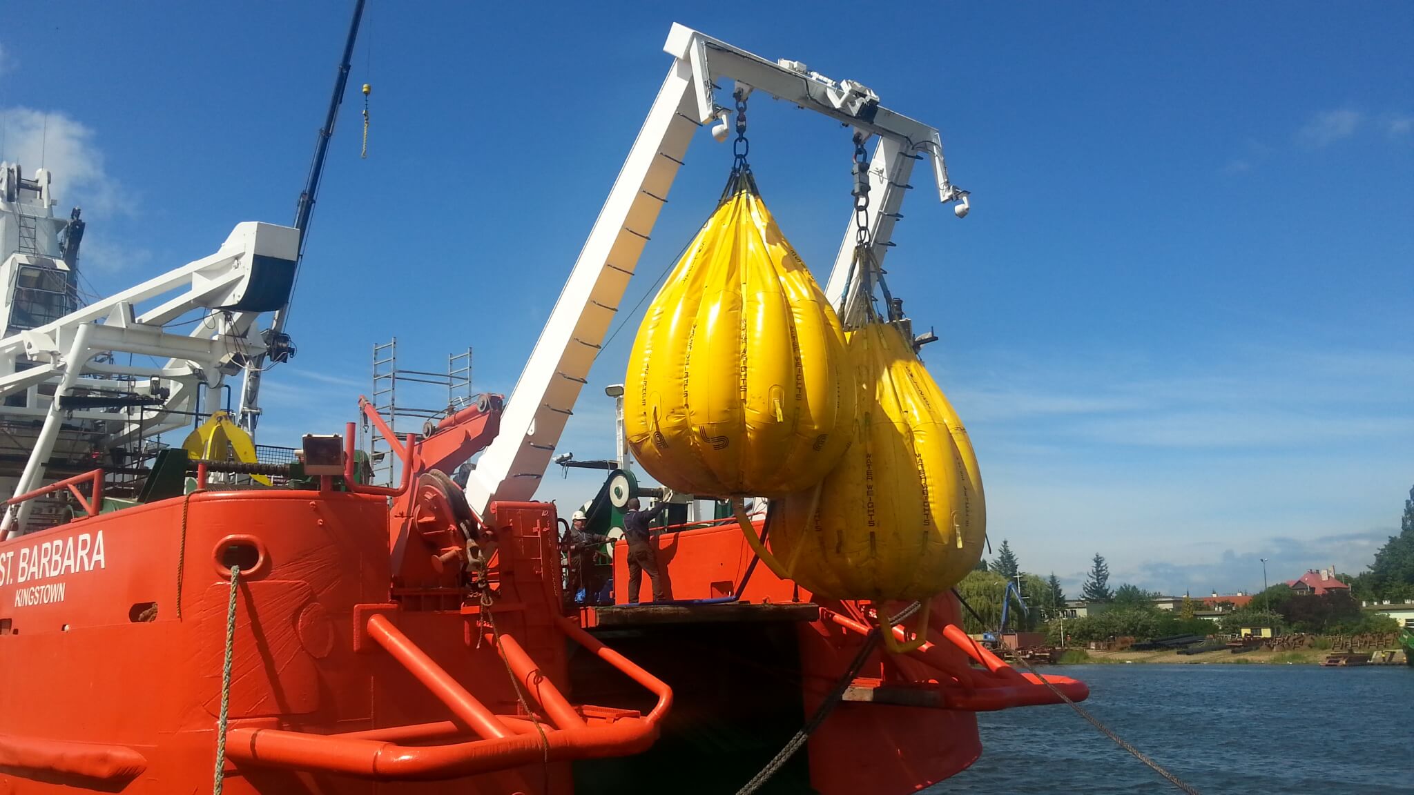 Na pierwszym planie widoczna jest masywna, czerwona jednostka o nazwie ST. BARBARA, zacumowana tuż przy brzegu. Z jej pokładu wznosi się biały wysięgnik dźwigowy, do którego przymocowano kilka dużych, żółtych worków wypełnionych wodą — typowe obciążniki wykorzystywane w testach wytrzymałościowych sprzętu podnoszącego. Ich zawieszenie w powietrzu sygnalizuje trwającą próbę obciążeniową, mającą na celu sprawdzenie nośności i bezpieczeństwa operacyjnego dźwigu.