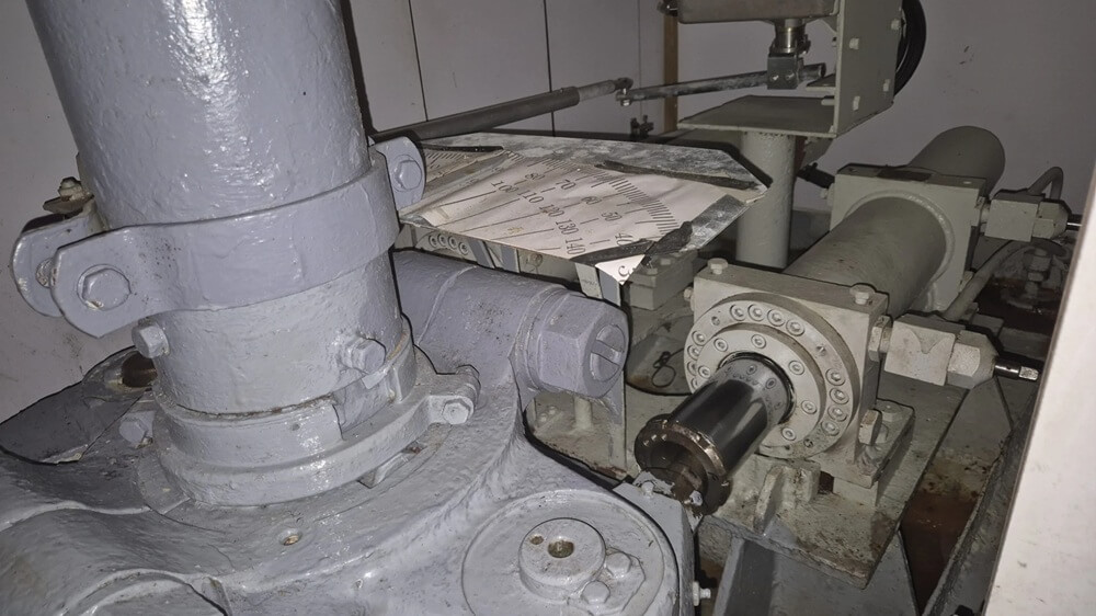 Na zdjęciu widoczny jest fragment maszyny przemysłowej — prawdopodobnie pompy lub zespołu napędowego — złożonej z cylindrycznych obudów, kołnierzy, śrub i połączeń rurowych. Całość utrzymana jest w odcieniach szarości, co podkreśla techniczny, surowy charakter urządzenia. Na jednym z elementów leży kartka z wydrukowanymi schematami i tekstem, co sugeruje trwające prace serwisowe lub diagnostyczne. Otoczenie wskazuje na wnętrze zakładu lub pomieszczenia technicznego.