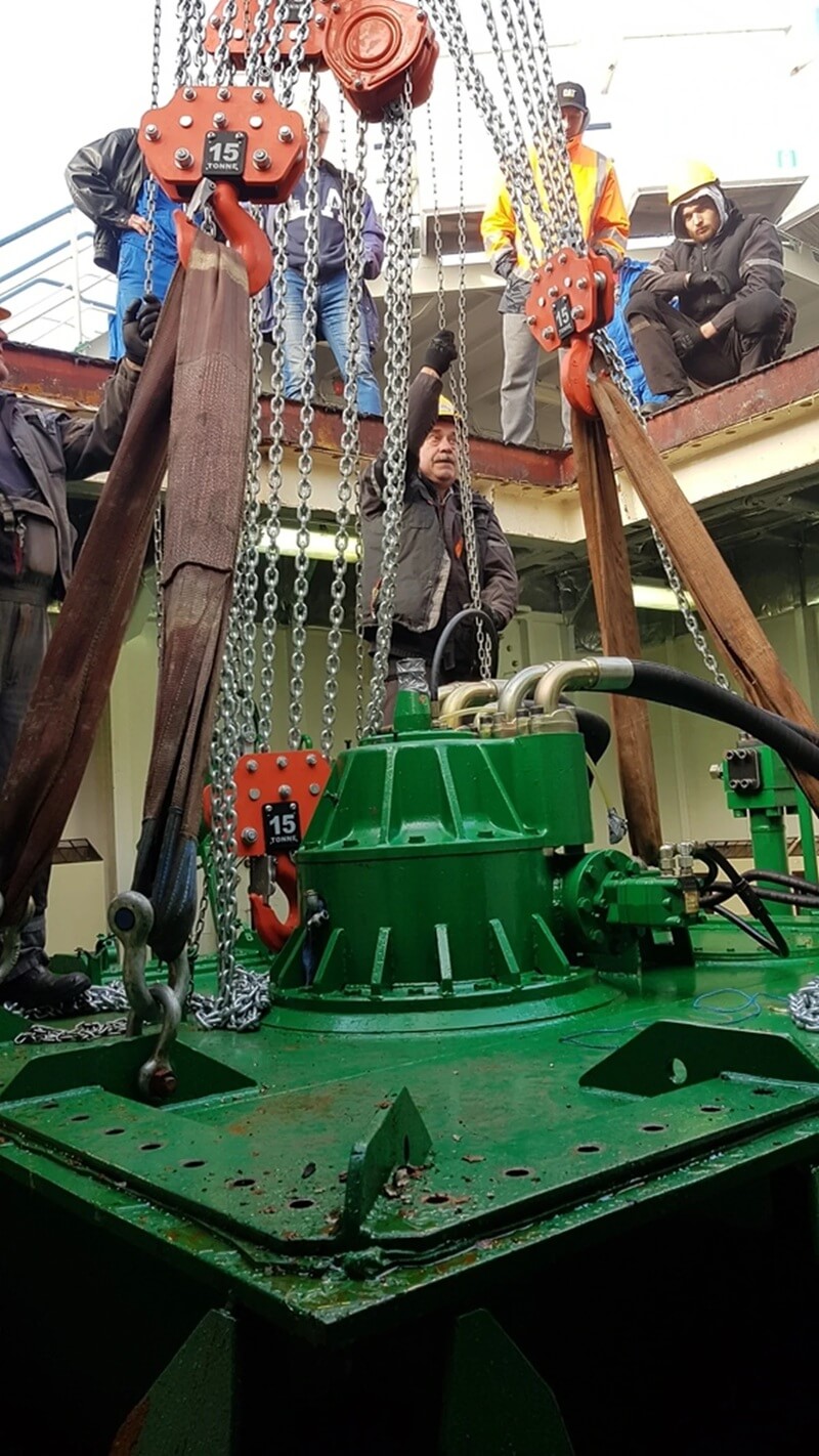 Na pokładzie platformy lub statku trwa precyzyjna operacja z udziałem dużego, zielonego urządzenia technicznego, zawieszonego na masywnych łańcuchach i pasach transportowych. Widoczne haki oznaczone „15T” wskazują na udźwig 15 ton, co podkreśla skalę i wagę obsługiwanego komponentu — prawdopodobnie elementu systemu hydraulicznego lub podwodnego. Kilku pracowników w odzieży roboczej koordynuje podnoszenie lub opuszczanie urządzenia przez otwór w pokładzie. Scena ukazuje złożoność pracy w środowisku morskim lub przemysłowym, gdzie liczy się siła, precyzja i zespołowa synchronizacja.