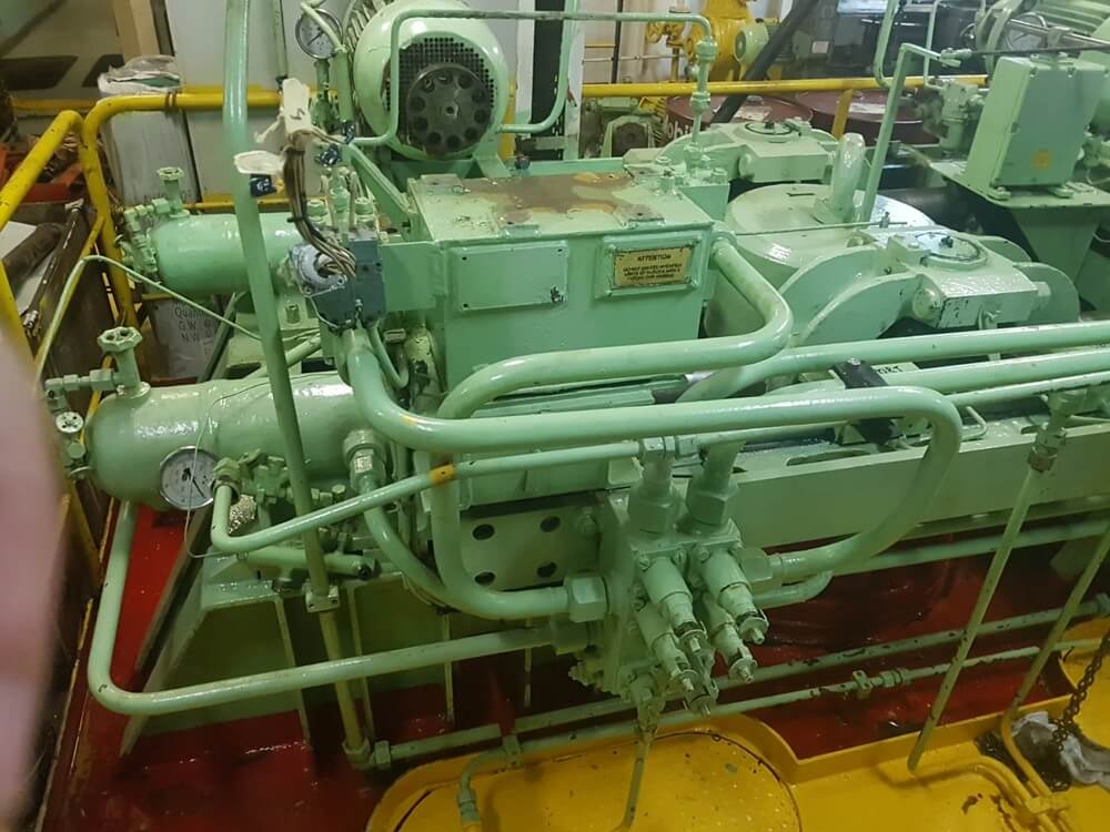 Na czerwono-żółtej platformie znajduje się duża maszyna sterowa — prawdopodobnie sprężarka lub układ hydrauliczny — pomalowana na jasnozielono. Widoczne są silnik, zawory, manometry, rury i jednostki sterujące, co wskazuje na złożony system obsługujący przepływ cieczy lub gazu. Całość otoczona jest żółtymi barierkami ochronnymi, co sugeruje, że urządzenie znajduje się w hali produkcyjnej lub pomieszczeniu technicznym. Tabliczka znamionowa na centralnym module może zawierać dane identyfikacyjne lub parametry techniczne.