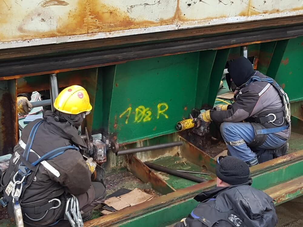 Trzech pracowników w odzieży ochronnej prowadzi działania montażowe lub serwisowe na dużej zielonej belce stalowej. Dwóch z nich używa elektronarzędzi do pracy przy śrubach lub mocowaniach, trzeci wspiera działania. Widoczne są kaski, rękawice i uprzęże, co świadczy o wysokim standardzie bezpieczeństwa. Żółte oznaczenie „1D2R” na belce może wskazywać na identyfikator techniczny lub strefę roboczą. Tło sugeruje, że prace odbywają się w ramach dużej infrastruktury — być może mostu lub konstrukcji nośnej.