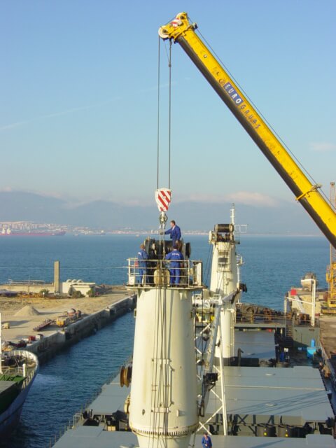 Na szczycie dużej cylindrycznej konstrukcji — prawdopodobnie części statku lub okrętu podwodnego — grupa pracowników w niebieskich kombinezonach i kaskach realizuje zadanie z użyciem dźwigu oznaczonego „EURO GRUAS”. Hak zawieszony nad nimi wskazuje na trwającą operację podnoszenia lub montażu ciężkiego komponentu. W tle widoczne są morze, portowe zabudowania i górzysty horyzont, co nadaje scenie przestrzenny, techniczno-morski charakter. Całość ukazuje złożoność i skalę pracy inżynieryjnej w środowisku stoczniowym.