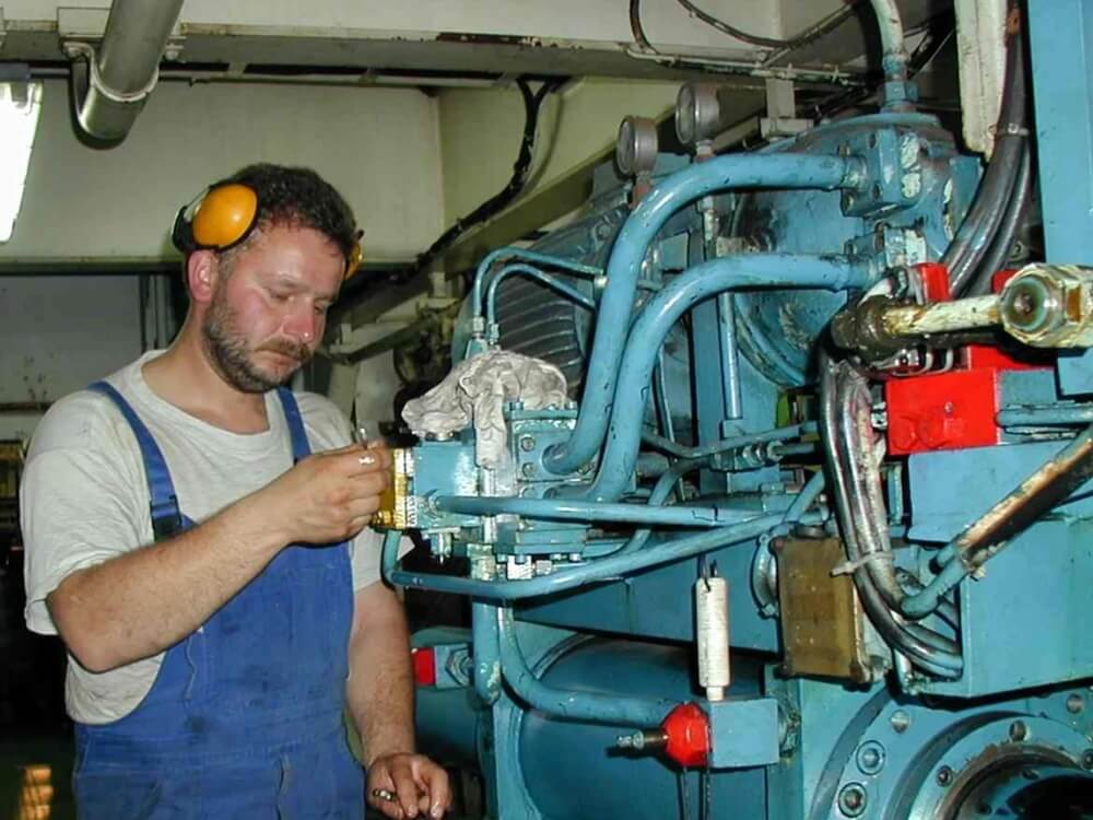 W przemysłowym otoczeniu mężczyzna w białej koszulce i niebieskich ogrodniczkach wykonuje czynności serwisowe przy dużej maszynie sterowej. Na uszach ma ochronniki słuchu, a w dłoni trzyma narzędzie, skupiając się na jednym z zaworów lub połączeń. Niebieska konstrukcja urządzenia, pełna rur i mechanicznych elementów, podkreśla złożoność obsługiwanej technologii. Scena emanuje spokojem, techniczną precyzją i rutyną zawodową.