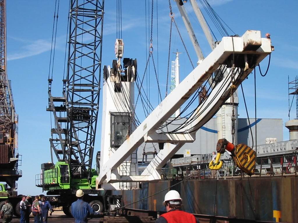 Na terenie stoczni lub dużego placu budowy trwa intensywna operacja z użyciem potężnego dźwigu o białym ramieniu i systemie czarnych lin nośnych. Zawieszony na nich jest masywny hak z żółto-czarnym oznaczeniem, gotowy do podniesienia ciężkiego ładunku. Na ziemi pracuje kilku techników w kaskach i kamizelkach odblaskowych, co świadczy o zachowaniu wysokich standardów BHP. W tle widać inne dźwigi, konstrukcje stalowe i statek, co podkreśla złożoność i skalę operacji logistyczno-technicznej.