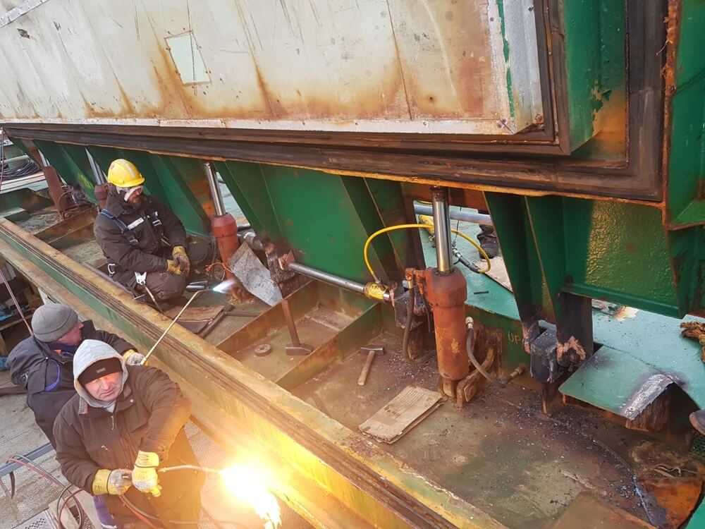 Na terenie zakładu przemysłowego grupa pracowników realizuje zadania związane z obróbką i montażem stalowych konstrukcji. Jeden z nich, na pierwszym planie, operuje palnikiem, generując intensywny płomień — prawdopodobnie do cięcia lub spawania. Pozostali obsługują narzędzia i elementy hydrauliczne, w tym siłownik i przewody podłączone do zielonej stalowej struktury. Wszyscy ubrani są w odzież ochronną: kaski, rękawice, kombinezony — co podkreśla wysoki poziom bezpieczeństwa w wymagającym środowisku pracy.