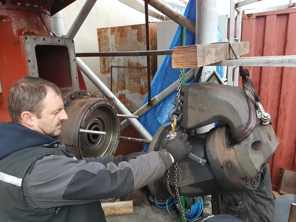 W otoczeniu przemysłowym mężczyzna w rękawicach roboczych obsługuje masywny element metalowy, zawieszony na łańcuchu i haku. Używa ręcznego podnośnika łańcuchowego, by precyzyjnie ustawić komponent — prawdopodobnie część silnika lub maszyny ciężkiej. W tle widoczne są inne elementy konstrukcyjne, rusztowanie i czerwony kontener, co sugeruje tymczasowe stanowisko naprawcze lub montażowe. Scena ukazuje fizyczny wymiar pracy technicznej, wymagającej siły, doświadczenia i dokładności.