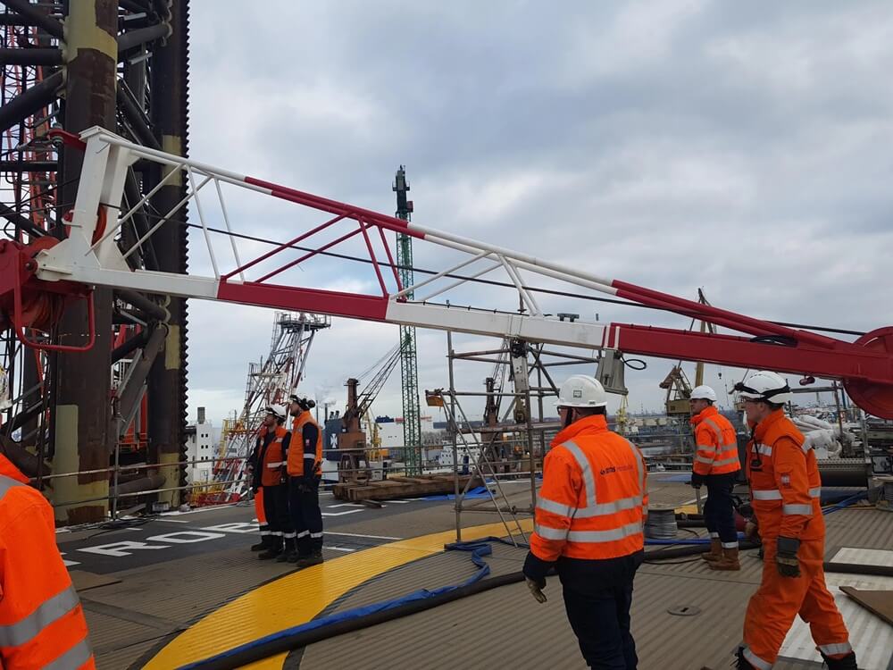 Na zdjęciu widać pracowników w odzieży ochronnej podczas prac na platformie przemysłowej/offshore. W centrum znajduje się duży żuraw z charakterystycznym czerwono-białym wysięgnikiem, a w tle widoczna jest infrastruktura portowa i przemysłowa. Pracownicy mają na sobie pomarańczową odzież ostrzegawczą, kaski i środki ochrony osobistej. Na platformie znajdują się również przewody, elementy wyposażenia technicznego oraz konstrukcje zabezpieczające.